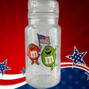 Pode incluir: Uma garrafa de vidro transparente com uma bandeira americana vermelha, branca e azul e dois personagens M&M's. Os personagens M&M's usam roupas vermelhas e verdes e seguram a bandeira.