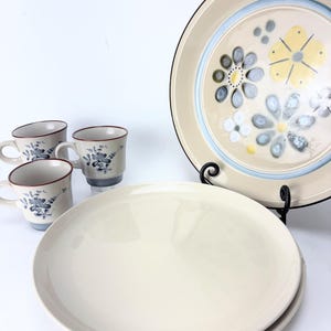 May include: A vintage cream-colored dinnerware set featuring four mugs with blue floral designs and a brown rim. The set includes two large cream-colored plates and a decorative plate with a floral pattern in yellow, blue, and gray.