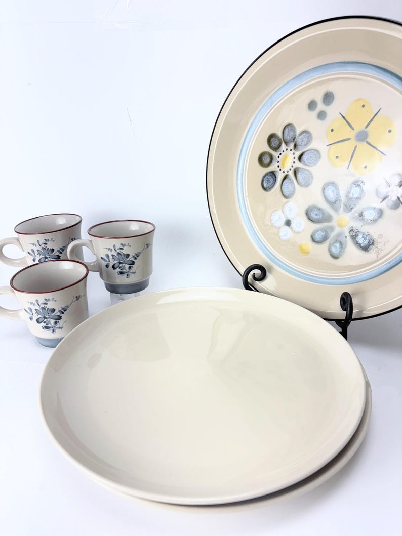 May include: A vintage cream-colored dinnerware set featuring four mugs with blue floral designs and a brown rim. The set includes two large cream-colored plates and a decorative plate with a floral pattern in yellow, blue, and gray.
