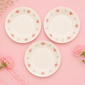May include: Three off-white ceramic plates with a pink heart and floral pattern border. The plates are arranged on a pink surface, with pink flowers and a pink lace cloth in the background.