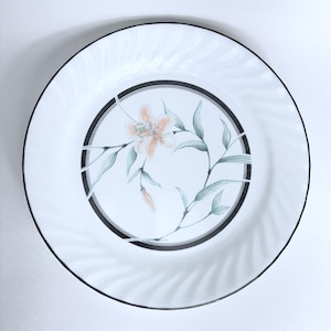 Puede incluir: Plato blanco con borde negro y diseño floral. El centro presenta una flor color melocotón con hojas y tallos verdes, enmarcada por un borde negro y gris. El borde del plato tiene un patrón sutil y texturizado.