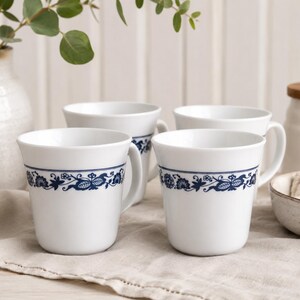 May include: Four white ceramic mugs with a blue floral design band. The mugs have a simple, rounded shape with a handle. The mugs are on a neutral-coloured cloth, with a vase and other kitchenware in the background.