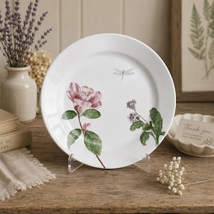 Pode incluir: Prato de cerâmica branco com ilustrações florais e de libélulas. O prato apresenta uma flor rosa, folhas verdes e um desenho de libélula. O prato é exibido em um suporte transparente.