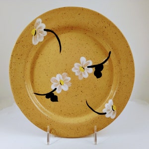 May include: A yellow ceramic plate with a speckled glaze features a design of white daisies with yellow centers and black stems. The daisies are arranged in a stylized pattern around the rim of the plate.