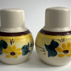 May include: A pair of ceramic salt and pepper shakers with a brown band and yellow and green floral designs. The shakers feature a sunflower design with brown centres.