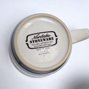 May include: A ceramic mug with a handle, viewed from the bottom. The mug is off-white with a gray band around the rim. The bottom features the text "Noritake STONEWARE" and "PLEASURE".