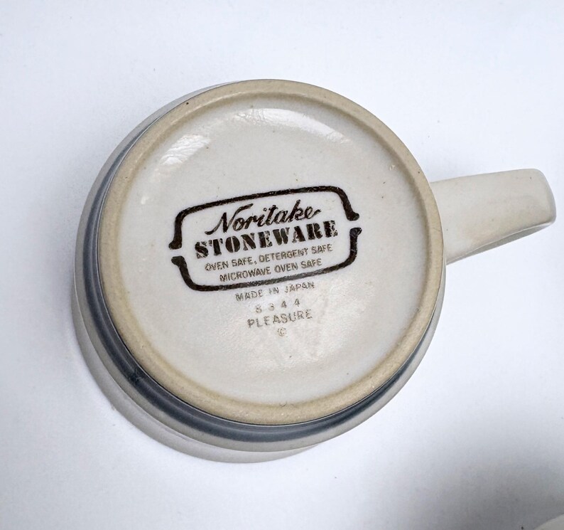 May include: A ceramic mug with a handle, viewed from the bottom. The mug is off-white with a gray band around the rim. The bottom features the text "Noritake STONEWARE" and "PLEASURE".
