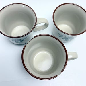 May include: Three ceramic mugs with a beige interior and exterior. Each mug has a brown rim and handle. The mugs have a floral design in teal. The mugs are arranged in a triangular pattern.