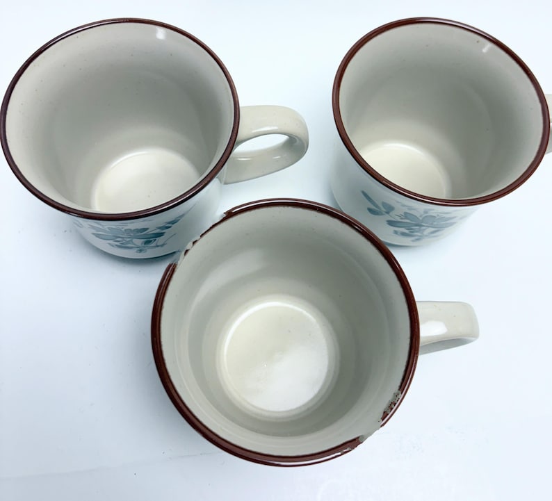 May include: Three ceramic mugs with a beige interior and exterior. Each mug has a brown rim and handle. The mugs have a floral design in teal. The mugs are arranged in a triangular pattern.