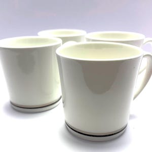 May include: Four white ceramic mugs with a cream-colored interior. Each mug has a handle and a decorative band of black and tan near the base. The mugs are arranged on a white surface.