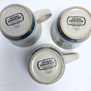 May include: Three Noritake stoneware mugs, viewed from the bottom, showcasing the brand's logo and the word "PLEASURE." The mugs have a light beige base with a blue floral design and a matching blue band near the rim. Each mug has a curved handle.