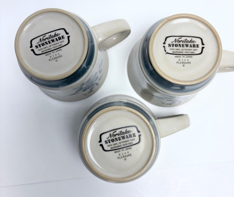 May include: Three Noritake stoneware mugs, viewed from the bottom, showcasing the brand's logo and the word "PLEASURE." The mugs have a light beige base with a blue floral design and a matching blue band near the rim. Each mug has a curved handle.