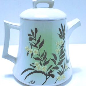 May include: A white ceramic teapot with a lid, handle, and spout. The teapot is decorated with a floral design featuring brown leaves and small white flowers against a light green gradient background. The handle is rectangular.