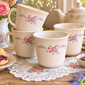 Pode incluir: Uma coleção de chávenas e canecas creme com um design floral. As chávenas e canecas são decoradas com rosas cor-de-rosa e detalhes azuis. Um prato com um biscoito e geleia, uma chaleira e um vaso de flores também são visíveis.