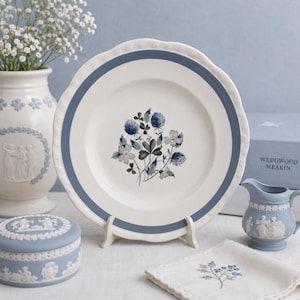 Puede incluir: Un plato blanco con borde azul y diseño floral, exhibido sobre un soporte blanco. El plato presenta un grupo de flores azules y grises. Otros artículos incluyen un jarrón, una caja pequeña y una jarra de crema, todos en una combinación de colores azul y blanco similar.