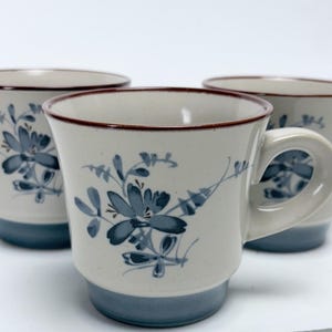 May include: Three ceramic mugs with a floral design. The mugs are off-white with blue floral patterns and a blue base. Each mug has a brown rim and a handle.