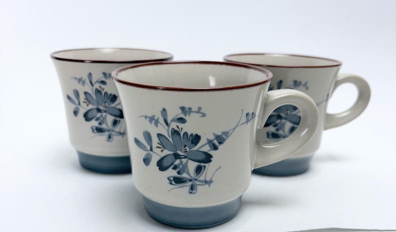 May include: Three ceramic mugs with a floral design. The mugs are off-white with blue floral patterns and a blue base. Each mug has a brown rim and a handle.