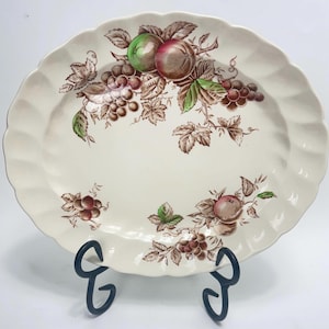 Puede incluir: Un plato ovalado blanco para servir con un diseño floral marrón y verde con uvas y manzanas. El plato tiene un borde festoneado y está sobre un soporte negro.