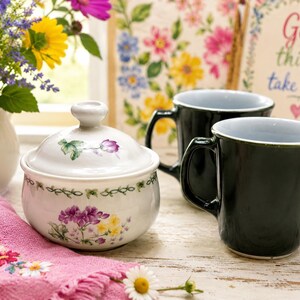 May include: A white ceramic sugar bowl with a floral design, a pink embroidered napkin, and two black mugs with gray interiors. A vase of flowers and a framed sign with the words "Good things take time" are in the background.