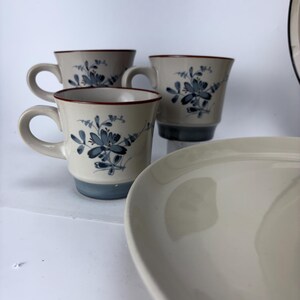 May include: A set of ceramic tableware featuring three mugs and a plate. The mugs have a beige base, blue floral design, and a red rim. The plate is off-white and has a curved shape. The set is on a white surface.