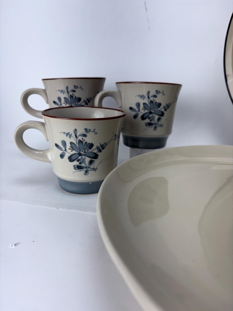 May include: A set of ceramic tableware featuring three mugs and a plate. The mugs have a beige base, blue floral design, and a red rim. The plate is off-white and has a curved shape. The set is on a white surface.