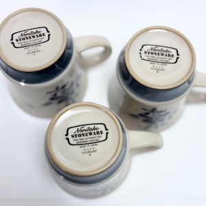 May include: Three Noritake stoneware mugs with handles, featuring a cream-colored base and a dark blue rim. The mugs have a floral design and the text "Noritake Stoneware" is visible on the bottom. The mugs are made in Japan.