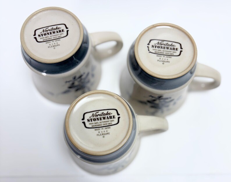 May include: Three Noritake stoneware mugs with handles, featuring a cream-colored base and a dark blue rim. The mugs have a floral design and the text "Noritake Stoneware" is visible on the bottom. The mugs are made in Japan.