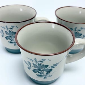 May include: Three white ceramic mugs with a floral design in blue and brown trim. The mugs have a light blue base and a curved handle. The floral design is repeated on each mug.
