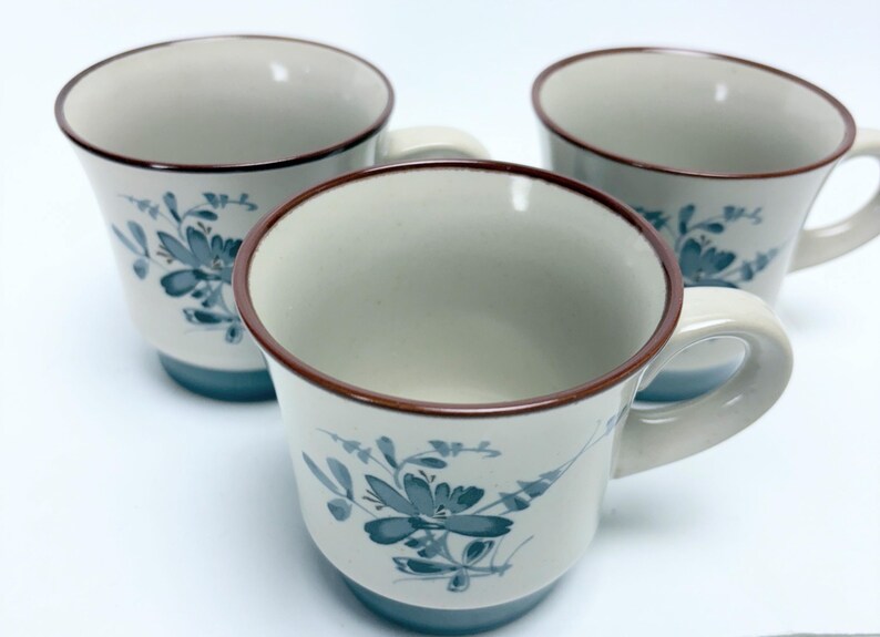 May include: Three white ceramic mugs with a floral design in blue and brown trim. The mugs have a light blue base and a curved handle. The floral design is repeated on each mug.
