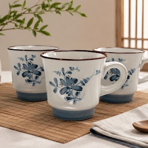May include: Three white ceramic mugs with blue floral designs and brown rims. The mugs have a blue base and white handles. A small vase with green leaves and a small dish are also in the image.