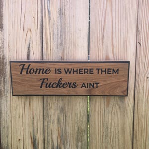 May include: Wooden sign with the phrase "Home is Where Them Fuckers Ain't" in black lettering. The sign has a dark border and is mounted on a wooden fence.