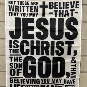 May include: White banner with black text. The banner reads "BUT THESE ARE WRITTEN THAT YOU MAY JESUS IS THE CHRIST, THE SON OF GOD, & THAT BELIEVING YOU MAY HAVE LIFE IN HIS NAME. JOHN 20:31". A cross is also featured.