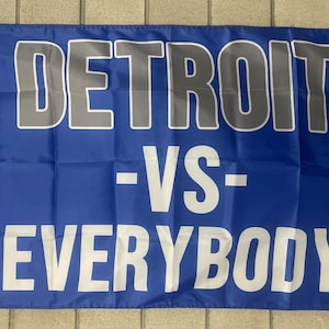 Puede incluir: Bandera azul con texto blanco y gris. La línea superior dice "DETROIT" en letras grises con contorno blanco. La segunda línea dice "-VS-" en blanco. La línea inferior dice "EVERYBODY" en blanco.