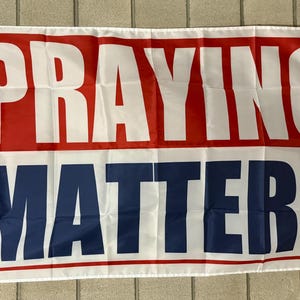 May include: White flag with the words "PRAYING" in large white letters on a red background, and "MATTERS" in white letters on a blue background. The flag has a white border and grommets.