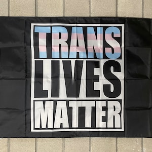 以下が含まれることがあります： 白文字で「Trans Lives Matter」と書いてある黒い旗。テキストは白い枠で囲まれています。テキストの上部は、トランスジェンダーのプライドフラッグに似ている、薄い青、ピンク、薄い青の色合いのパターンになっています。