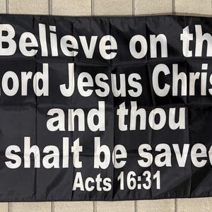 May include: Black flag with white text that reads "Believe on the Lord Jesus Christ, and thou shalt be saved" with "Acts 16:31" below. The flag has a white border and grommets for hanging.
