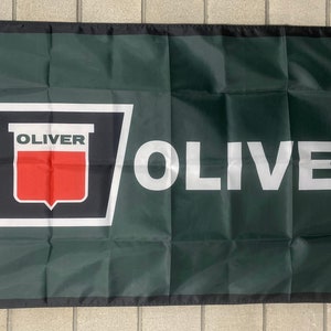 Puede incluir: Una bandera verde con un borde blanco y la palabra "OLIVER" en letras blancas. Un logotipo blanco y negro con la palabra "OLIVER" en letras rojas está en el lado izquierdo de la bandera.