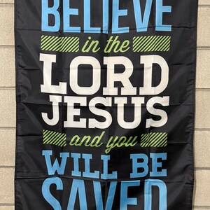 May include: Black banner with the words "BELIEVE in the LORD JESUS and you WILL BE SAVED" in white and blue lettering. The banner has green and white diagonal stripes. The banner is hanging on a brick wall.