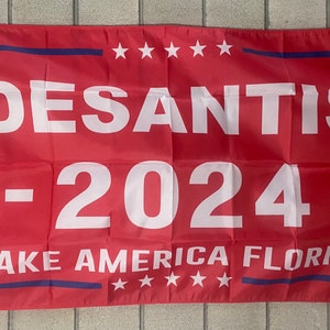 以下が含まれることがあります： 白の縁取りと青い星が付いた赤い旗。旗には「DESANTIS - 2024 - MAKE AMERICA FLORIDA」という文字が白で印刷されています。