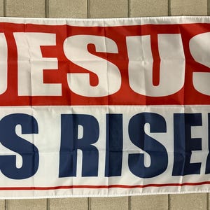 May include: A rectangular banner with the words "JESUS IS RISEN" in large white letters. The top section is red, and the bottom section is white with blue text. The banner has grommets for hanging.