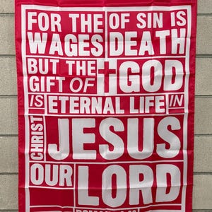 May include: Red flag with white text. The text reads: "FOR THE WAGES OF SIN IS DEATH BUT THE GIFT OF GOD IS ETERNAL LIFE IN CHRIST JESUS OUR LORD ROMANS 6:23". The flag has a white border and is hanging on a wall.