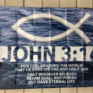 May include: A rectangular flag featuring a blue water wave background with a white Christian fish symbol. The text "JOHN 3:16" is prominently displayed, along with a Bible verse. The flag has a white border and grommets.
