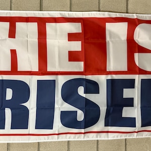 May include: A rectangular flag with the words "HE IS RISEN" in bold, white letters. The top section is red with white letters, and the bottom section is blue with white letters. The flag has a white border and grommets.