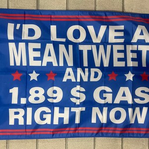 May include: A blue flag with white text that reads "I'd love a mean tweet and 1.89 $ gas right now". There are three red stars on each side of the word "and".