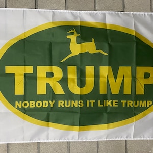May include: A white flag with a green and yellow oval. Inside the oval is a yellow deer with antlers and the text "TRUMP" in yellow letters. Below the oval is the text "NOBODY RUNS IT LIKE TRUMP".
