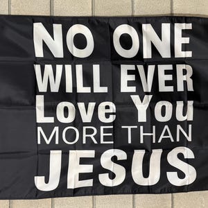 May include: Black flag with white text that reads "NO ONE WILL EVER Love You MORE THAN JESUS". The flag has a white border and grommets for hanging. The flag is made of a smooth, dark material.