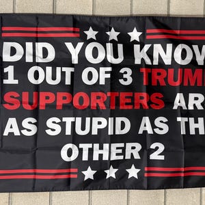 May include: Black flag with white text that reads "DID YOU KNOW 1 OUT OF 3 TRUMP SUPPORTERS ARE AS STUPID AS THE OTHER 2." The word "TRUMP" and "SUPPORTERS" are in red. The flag has three white stars at the top and bottom.