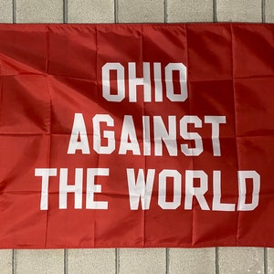 May include: A red flag with white text that reads "OHIO AGAINST THE WORLD." The flag has a white border and grommets for hanging. The flag is rectangular and made of a lightweight material.