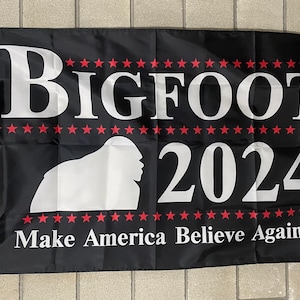 May include: Black and white flag with the text "BIGFOOT 2024" in white letters. The flag also features a white silhouette of Bigfoot and the text "Make America Believe Again!" in white letters. The flag has a red, white, and blue color scheme.