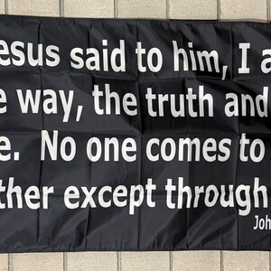 May include: Black flag with white text that reads "Jesus said to him, I am the way, the truth and the life. No one comes to the Father except through me. John 14:6". The flag has a white border and grommets.